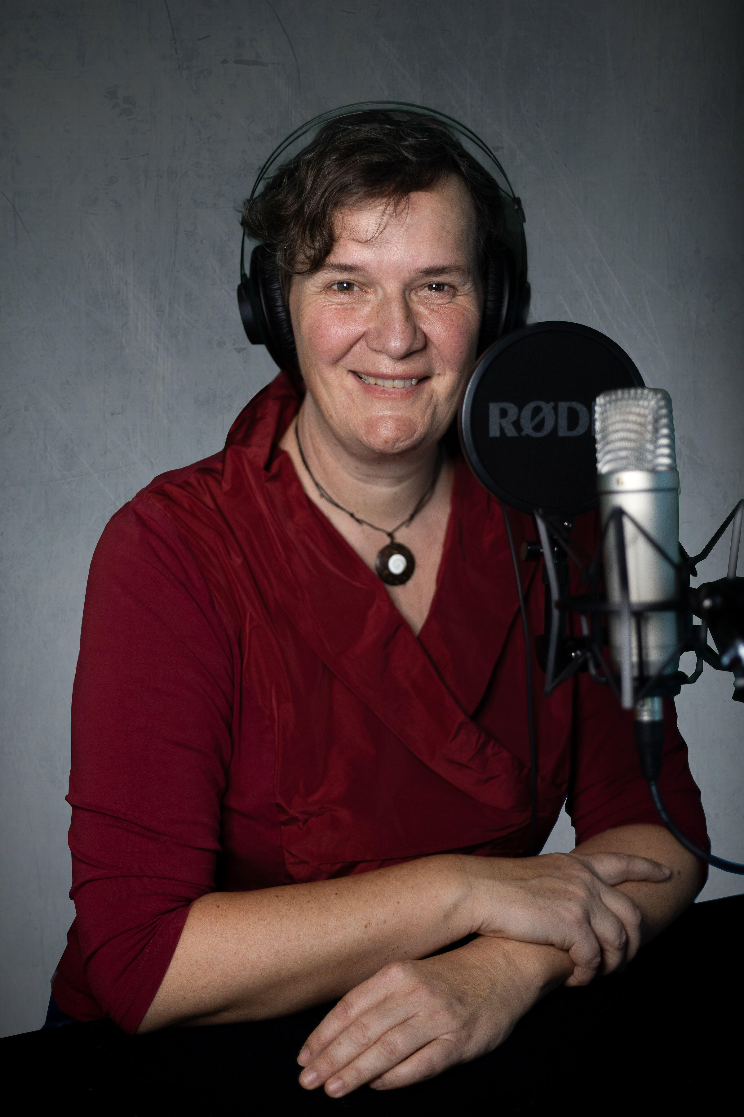 Profilfoto von Johanna Bahurai in roter Bluse mit Kopfhörer auf dem Kopf vor einem Studio-Mikrofon, in die Kamera lächelnd.